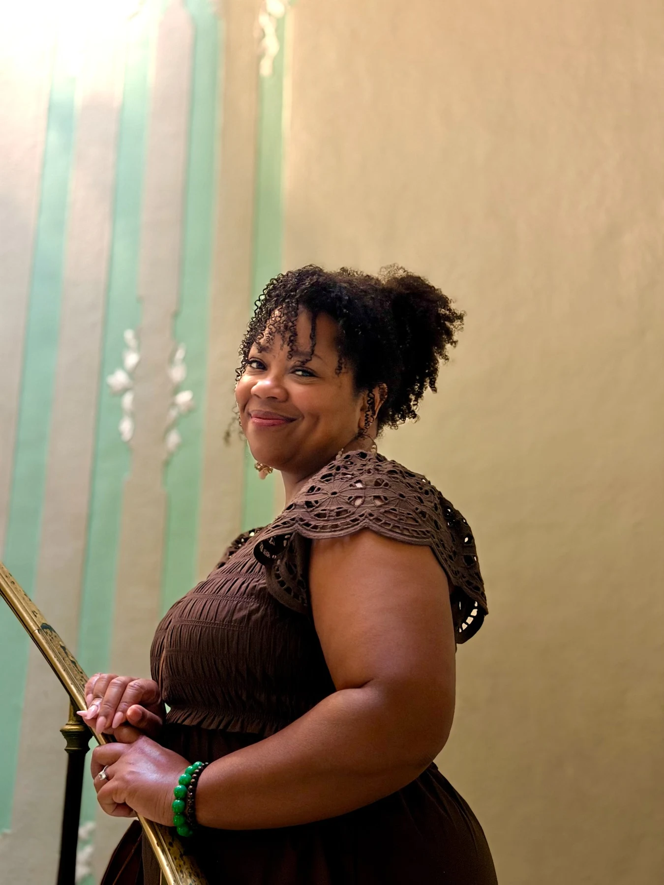 Shay smiling beside a staircase railing in a brown dress